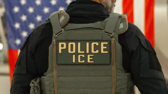 Umukozi ushinzwe umutekano wo kugenzura abimukira yafotorewe mu bitugu, yambaye ijaketi irinda amasasu irimo amagambo “police” na “ICE”
