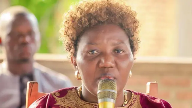Rose Nkorerimana arimo kuvugira mu ndangururamajwi. Yambaye ikanzu iboneka igice cyo hejuru iriho ibara ritukura na khaki. Inyuma ye hari umugabo utaboneka neza.