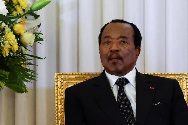 Perezida Paul Biya yicaye mu intebe areba muri camera. Yambaye ikote ry'umukara n'ishati y'umweru mo imbere na karuvati y'umukara irimo utudomo tw'umweru