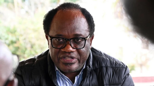 Umukandida w’abatavuga rumwe n’ubutegetsi, Brian Mundubile yambaye ikote ry'umukara. Mo imbere, harimo ishati irimo imirongo y'umweru n'ubururu