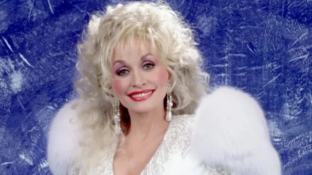 Dolly Parton yambaye imyenda y'umweru n'amaherena y'umweru. Ari guseka areba muri 'camera', inyuma ye hasa n'ubururu burimo uturongo dukobaguritse tw'umweru.