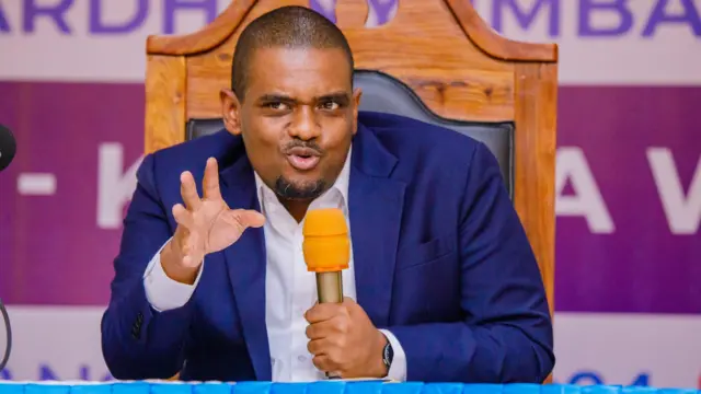 Deogratius Ndejembi akoze ikimenyetso n'ikiganza, yicaye arimo kuvugira mu ndangururamajwi iriho ipamba ry'ibara rya 'orange'. Yambaye ikositimu y'ubururu n'ishati y'umweru. Yicaye ku ntebe yegamirwa ikoze mu giti iriho umusego w'umukara.