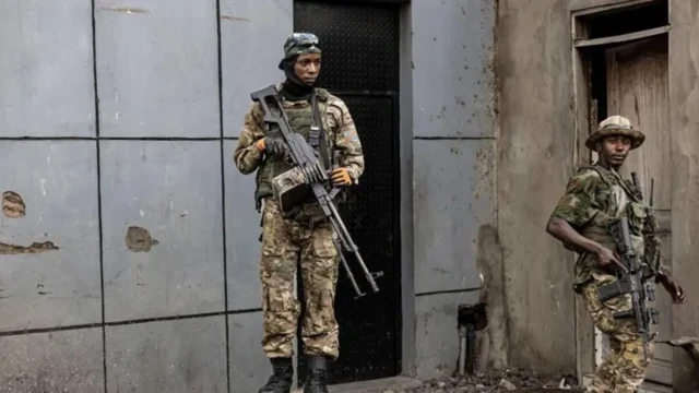 Abagabo babiri bo mu nyeshyamba za M23 bambaye imyenda ya gisirikare bahagaze bafite imbunda nini, i Goma, mu burasirazuba bwa Repubulika ya Demokarasi ya Congo, ku itariki ya 23 Gashyantare (2) mu 2025. Inyuma yabo hari inkuta z'inzu.