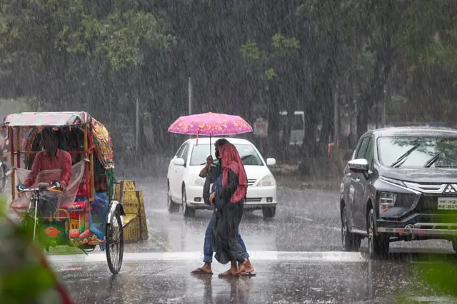 বৃষ্টির মধ্যে ছাতা মাথায় হেঁটে যাচ্ছেন দুইজন। একপাশে দুটি প্রাইভেটকার ও অন্যপাশে একটি রিকশা