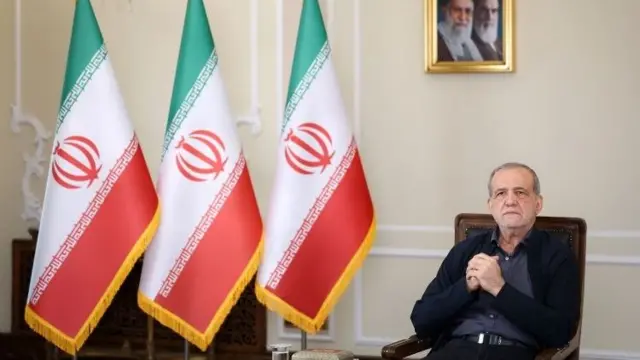 مسعود پزشکیان نشسته روی صندلی در کنار سه پرچم جمهوری اسلامی ایران