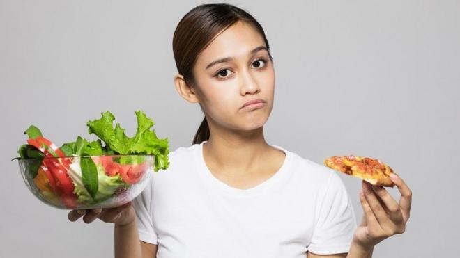 Una mujer joven sosteniendo una ensalada en una mano y en la otra un trozo de pizza.