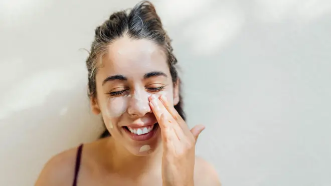 Retrato en primer plano de una joven aplicándose crema en las mejillas y la frente.