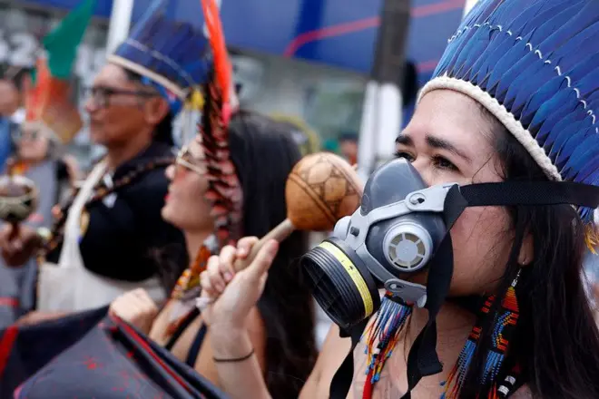 Mulher indígena usando cocar e uma máscara de gás, enquanto segura um chocalho com a mão direita, durante a Marcha Global pelo Clima, evento paralelo à COP30, em Belém. Ao lado dela estão outras pessoas indígenas com adereços tradicionais