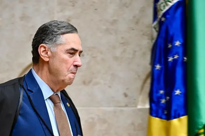 Barroso caminhando, com olhar confiante e leve sorriso, no STF; atrás dele, uma bandeira do Brasil