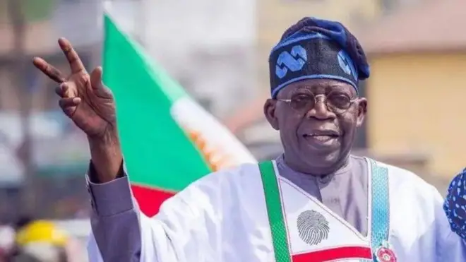 Aare Bola Tinubu na ika ọwọ ẹ meji soke, o n rẹrin-in. O wọ agbada funfun to ni itẹnu alawọ eeru, o de fila o si lo igo si oju rẹ