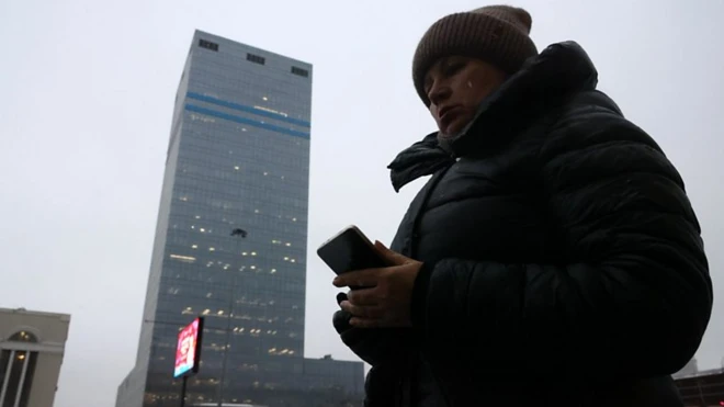 تصویری از نمای پایین یک زن که به گوشی موبایلش نگاه میکند و کلاه بافتنی به سر دارد، در کنار او یک ساختمان بلند دیده میشود