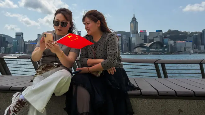 香港尖沙咀海旁兩位女士背對維多利亞港與香港島海岸線風景就坐於長椅上查看智能手機,其中一人手握一面小型中國國旗(18/3/2026)