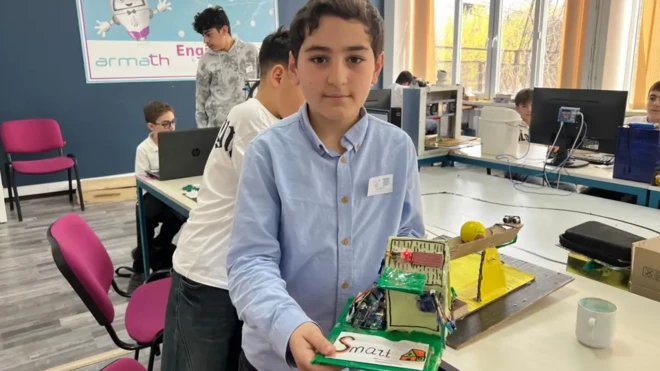 یک کودک در کلاس درس وسیلهای که ساخته را در دست گرفته در برابر دوربین نشان میدهد، چند دانشآموز دیگر در حال کار با کامپیوتر هستند