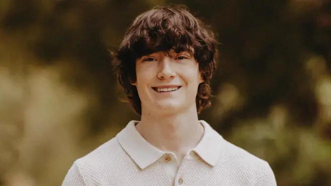 Фотографія Адама Рейна. У нього каштанове хвилясте волосся середньої довжини. Він посміхається, одягнений у в'язану сорочку з коміром і ґудзиками зверху. Позаду нього розмите тло з листям.