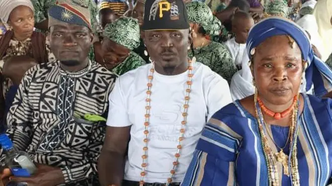 Ọkunrin kan to wọ aṣọ ankara dudu pẹlu fila jokoo lẹgbẹẹ arakunrin to wọ ẹwu funfun ati fila pẹlu ilẹkẹ lọrun, nigba ti iya kan naa wọ aṣọ ofi