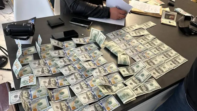 На столі розкладено велику кількість купюр по 100 доларів США, частина складена рівними рядами, інша — хаотично. Поруч видно документи, телефони, гаманець і пристрій для перевірки банкнот, а одна людина тримає аркуші паперу.
