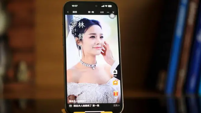 一张摆拍照片显示，一部手机直立放置，屏幕上播放一幕中国微短剧场景，一名新娘微笑着，新郎则抚摸她的脸。