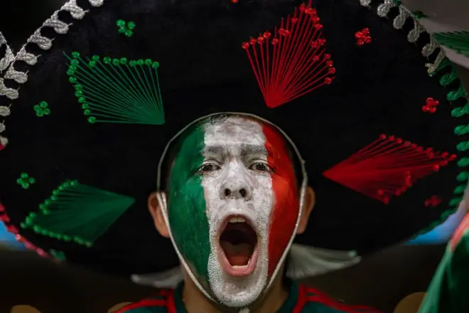 Un hombre con la boca abierta, seguramente está gritando o cantando. Tiene la cara pintada con los colores verde, blanco y rojo, que conforman la bandera de México. Lleva puesto un sombrero de mariachi.