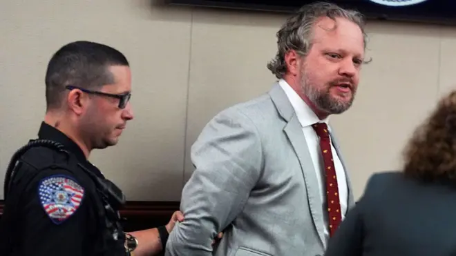 Джеймс Крейг, одягнений у сірий костюм, здається, розмовляє зі своїм адвокатом, поки поліцейський ліворуч тримає його за руку та відводить