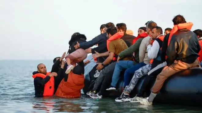 Множество людей в спасательных жилетах сидят на чем-то вроде надувного плота, трое из них стоят в воде, им с плота передают ребенка.