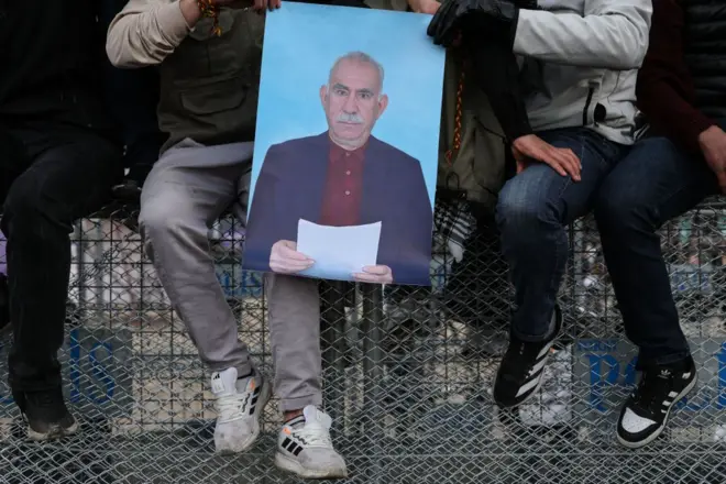 Polis barikatını üzerine oturan dört erkek. Yüzleri gözükmüyor. Ellerine Abdullah Öcalan posteri var. 