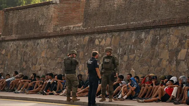 Soldados espanhóis conversam com um policial local enquanto observam migrantes sentados no chão, antes de escoltá-los de volta ao Marrocos, no enclave espanhol de Ceuta, no norte da África, em 1º de agosto de 2026. , no norte da África, informou hoje o Ministro do Interior da Espanha. 