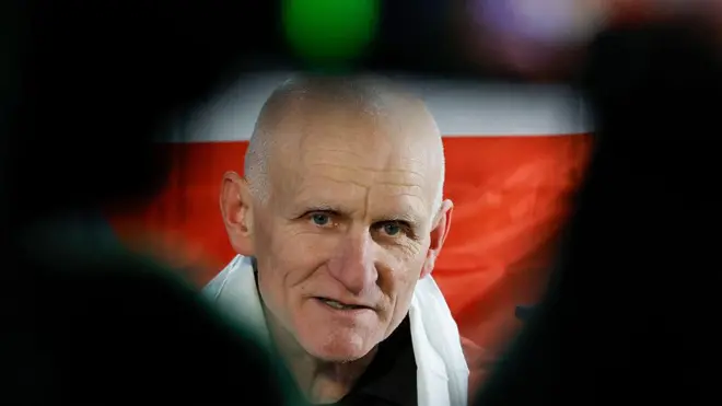 Крупным планом лицо Алеся Беляцкого на фоне бело-красно-белого флага белорусской оппозиции. При этом фотография обрамлена чем-то темным, снятым с очень близкого расстояния