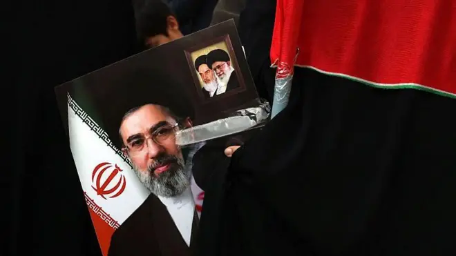 یکی از معترضان عکسی از آیت‌الله مجتبی خامنه‌ای، رهبر جدید جمهوری اسلامی ایران، در دست دارد؛ در کنار آن، تصاویر پدرش آیت‌الله علی خامنه‌ای، رهبر پیشین ایران، و آیت‌الله روح‌الله خمینی، رهبر معنوی جمهوری اسلامی نیز دیده می‌شود.