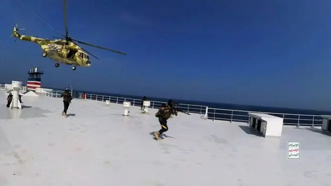 Un hélicoptère survole le pont d'un grand navire tandis que des hommes armés courent vers l'avant.