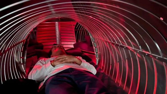 A man dressed in a white shirt lies on his back in a tunnel of red light for waht looks like some kind of experiment.