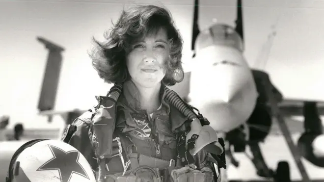 Donanma taktik uçaklarını kullanan ilk kadınlardan biri olan Teğmen Tammie Jo Shults, bir F/A-18'in önünde poz veriyor. Pilot tulumu giymiş ve kaskını sağ elinde tutuyor.