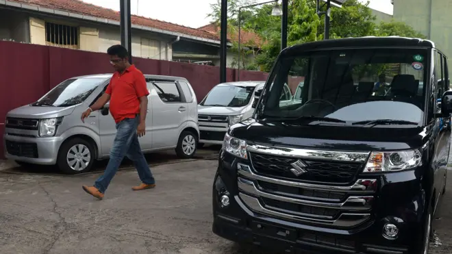 වාහන සේල් එකක නවතා ඇති අළු පැහැති වැගන් ආර් රථයක් සහ තවත් සුසුකි මෝටර් රථයක් පසු කරමින් පුද්ගලයෙකු ඇවිද යයි
