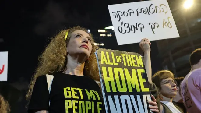 A woman stands at a protest holding a placard reading "All of them home now"