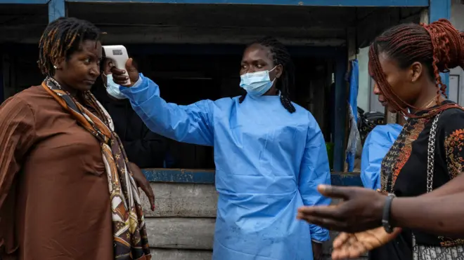Em uma rua na cidade de Goma, na República Democrática do Congo, um profissional de saúde com máscara facial azul e avental médico mede a temperatura de uma mulher com um dispositivo próximo à cabeça. Outra mulher e um homem estão do outro lado esperando. 