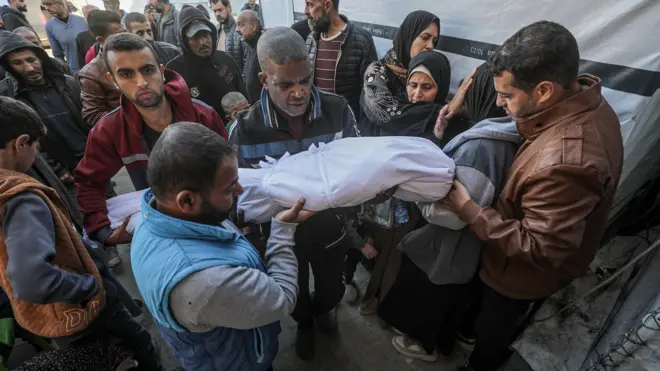 أب فلسطيني (وسط الصورة) يحمل جثمان ابنه شادي (14 عامًا) خلال جنازته في مستشفى الأقصى في دير البلح، وسط قطاع غزة.