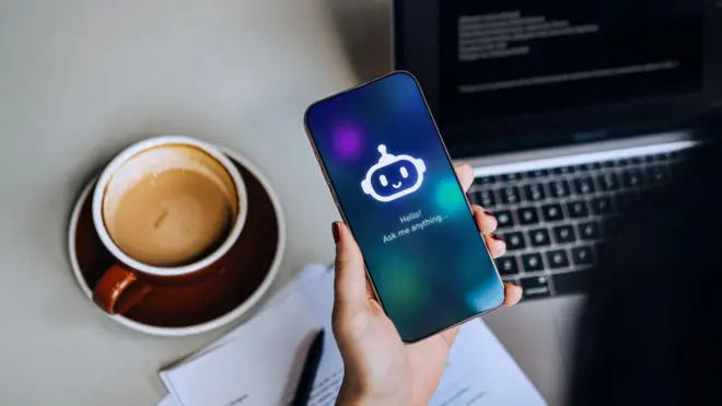 صورة من أعلى ليد سيدة تجلس أمام لابتوب، وأمامها كوب من القهوة، وتمسك بهاتف ذكي يظهر عليه صورة روبوت دردشة كتبت تحتها: Hello! Ask me anything.. 