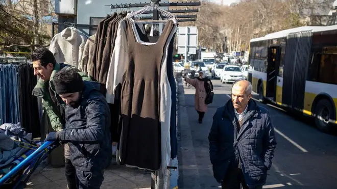 در یک خیابان دستفروشی پوشاک برای فروش گذاشته و چند عابر در حال عبور و مرور هستند