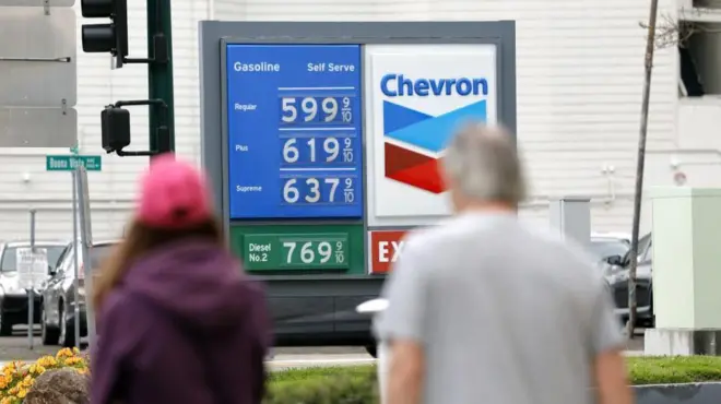 ガソリンスタンドに表示されている価格を、歩道から男女が見ている