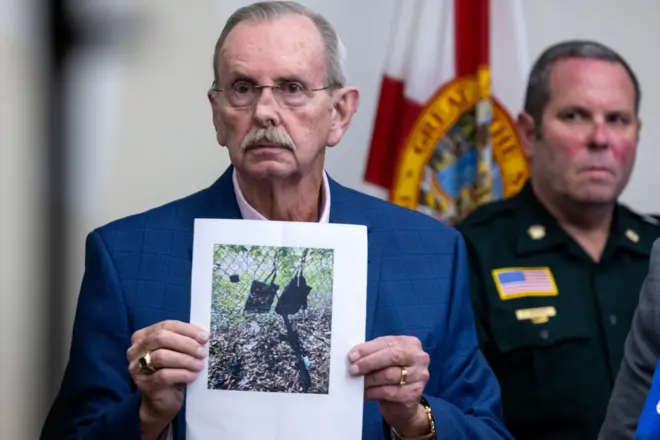 Homem idoso de terno azul marinho segura página com fotografia impressa, que mostra duas bolsas e um rifle