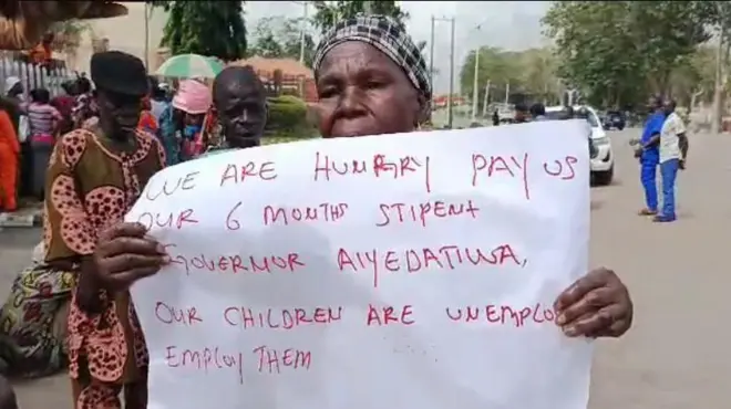 Iya agbalagba kan lara awon olufẹhonuhan naa gbe akọle to n kede pe Gomina Aiyedatiwa jẹ wọn lowo oṣu mẹfa dani