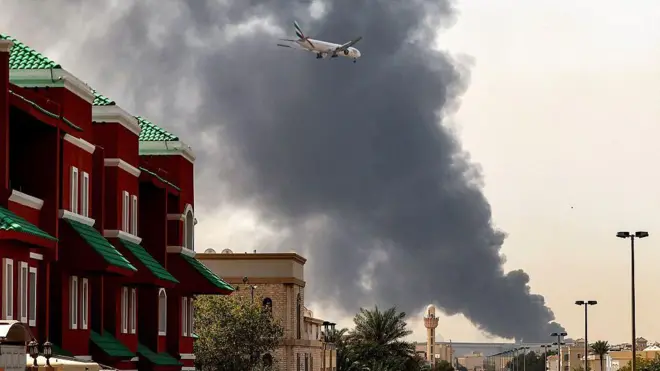 Малоэтажные жилые дома на фоне поднимащегося вдалеке стольа дыма: в небе виде пасажирский самолет на малой высоте