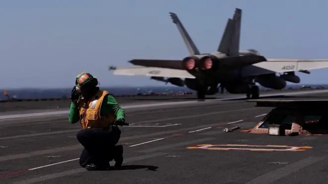 A man kneels down on the deck of aircraft carrier USS Gerald R Ford as a fighter plane is seen behind him (09/03/26)