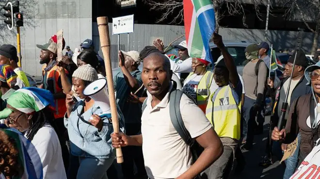 man hold stick while protesting, South Africa flag dey show for di side