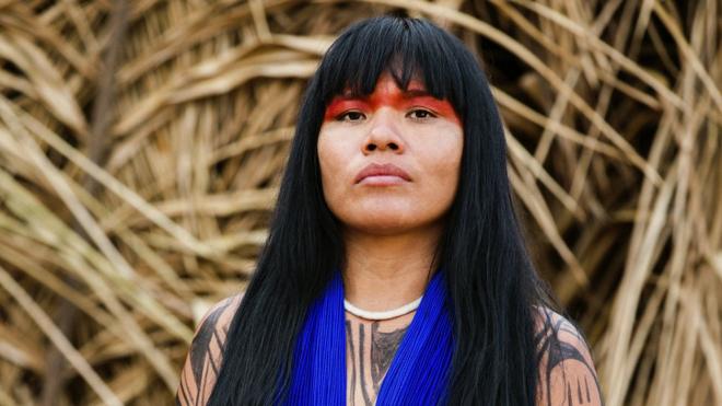 Fotografia colorida mostra uma mulher do povo Yawalapiti, com cabelos lisos e negros, usando uma pintura ritual vermelha e colaragenda de apostas on line da big senamiçangas azuis