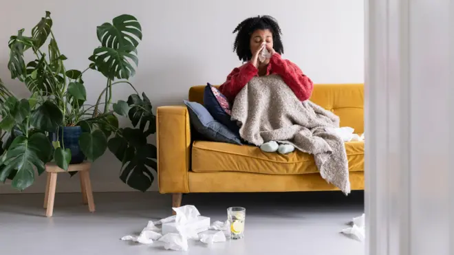 Uma pessoa sentada em um sofá amarelo, usando uma blusa vermelha, debaixo das cobertas assoa o nariz. No chão estão lenços de papel e um copo de água com uma rodela de limão.