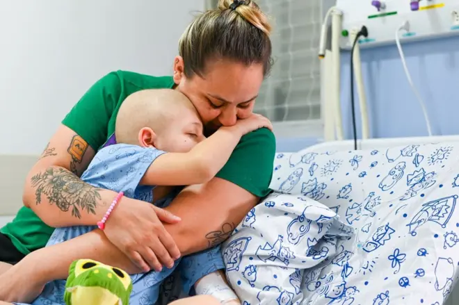 Mãe e filho abraçados na maca; menino não tem cabelo