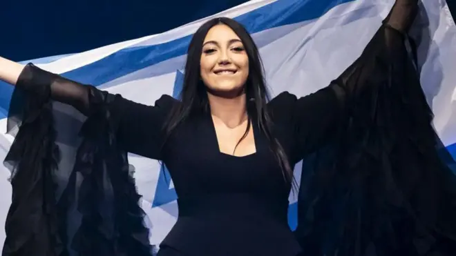  يوفال رافاييل ترتدي فستاناً أسود بأكمام طويلة وتحمل علم إسرائيل.