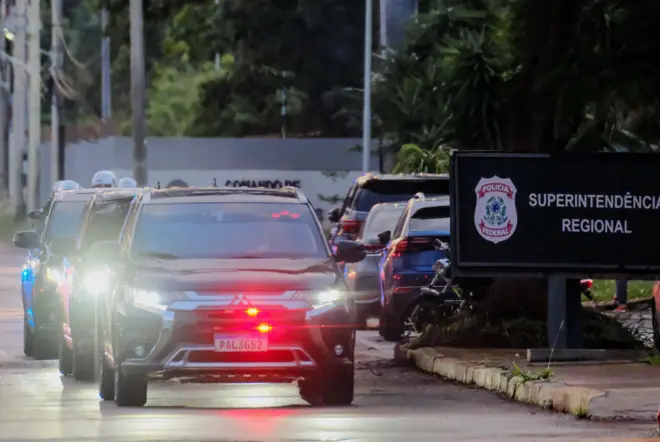Viaturas se movimentam na rua ao lado de placa da Polícia Federal