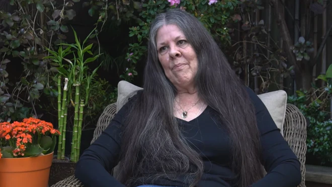 Loreen Willemberg, uma mulher de meia idade e cabelos longos, veste jeans e camisa de manga comprida preta 