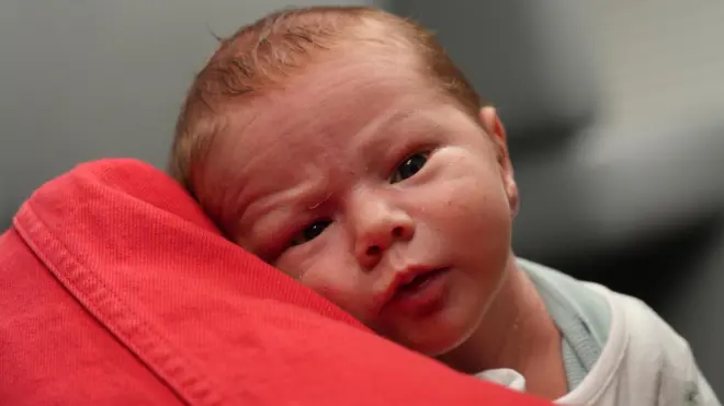 Baby Henry wide awake looking directly at the camera. He is resting on the arm of his grandmother who is wearing a red denim jacket. You can only see her arm. Henry is wearing a vest and a cream bodysuit. He has a tiny wrinkled forehead and is looking intruiged